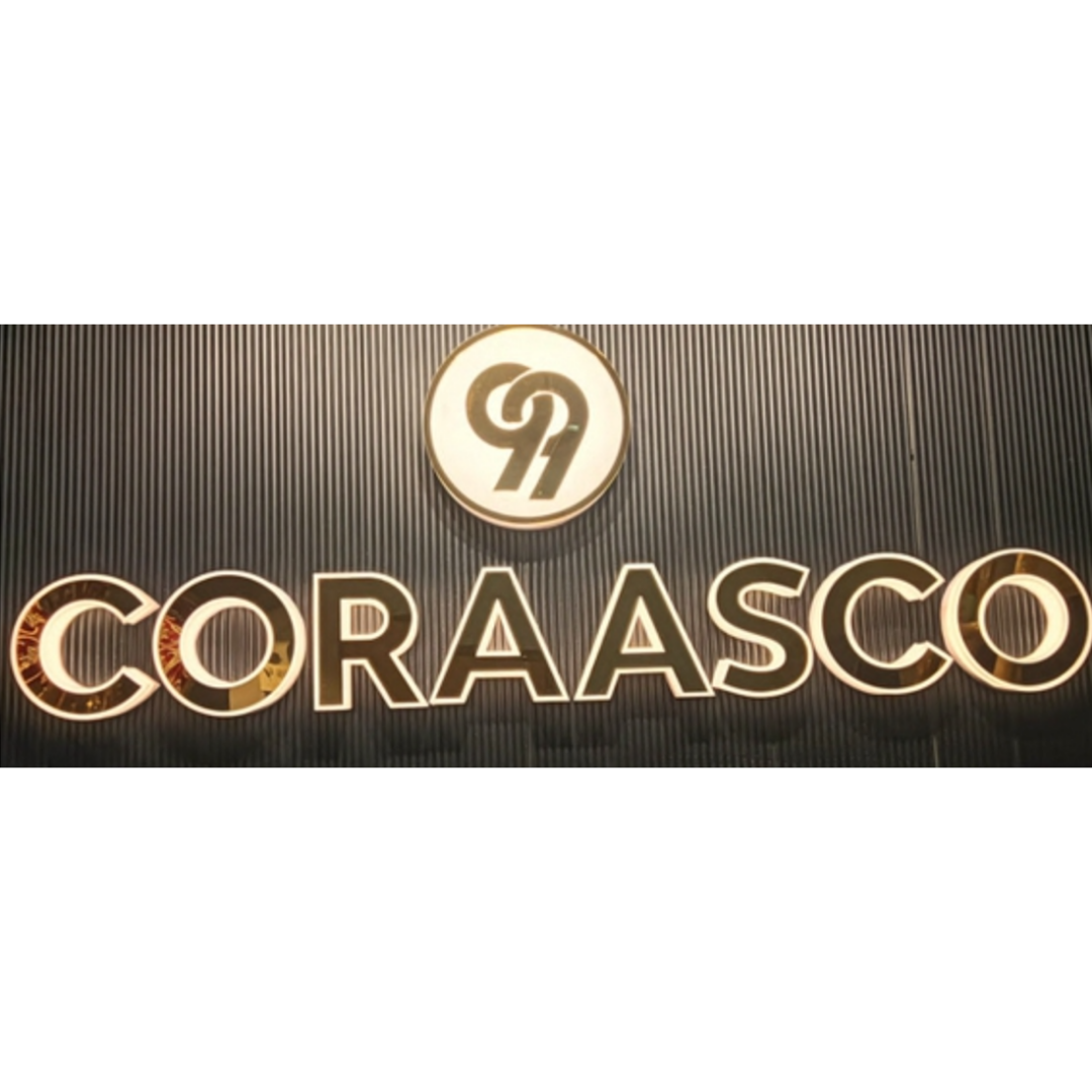 Large illuminated “CORAASCO” lettering with a circular logo above it on a dark vertically textured wall.