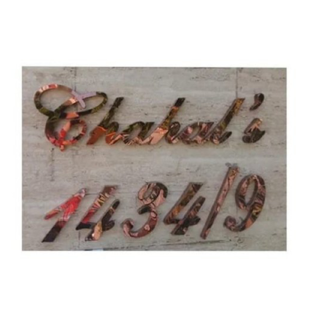 Close-up of a decorative wall sign featuring large raised cursive lettering in a metallic multicolor finish, with additional raised characters beneath it.