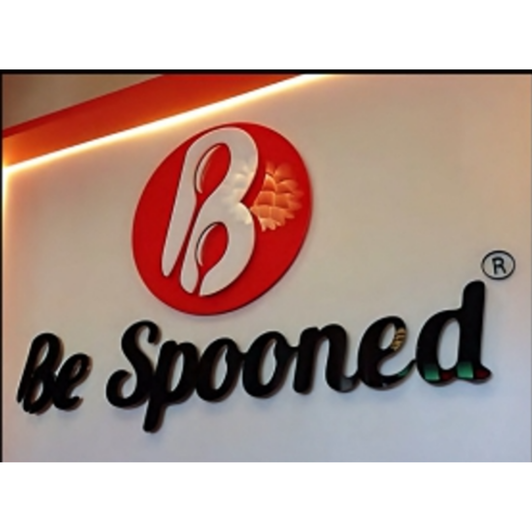 A wall-mounted “be spoonfed” logo sign featuring black raised lettering, a red circular emblem, and a stylized white letter B.